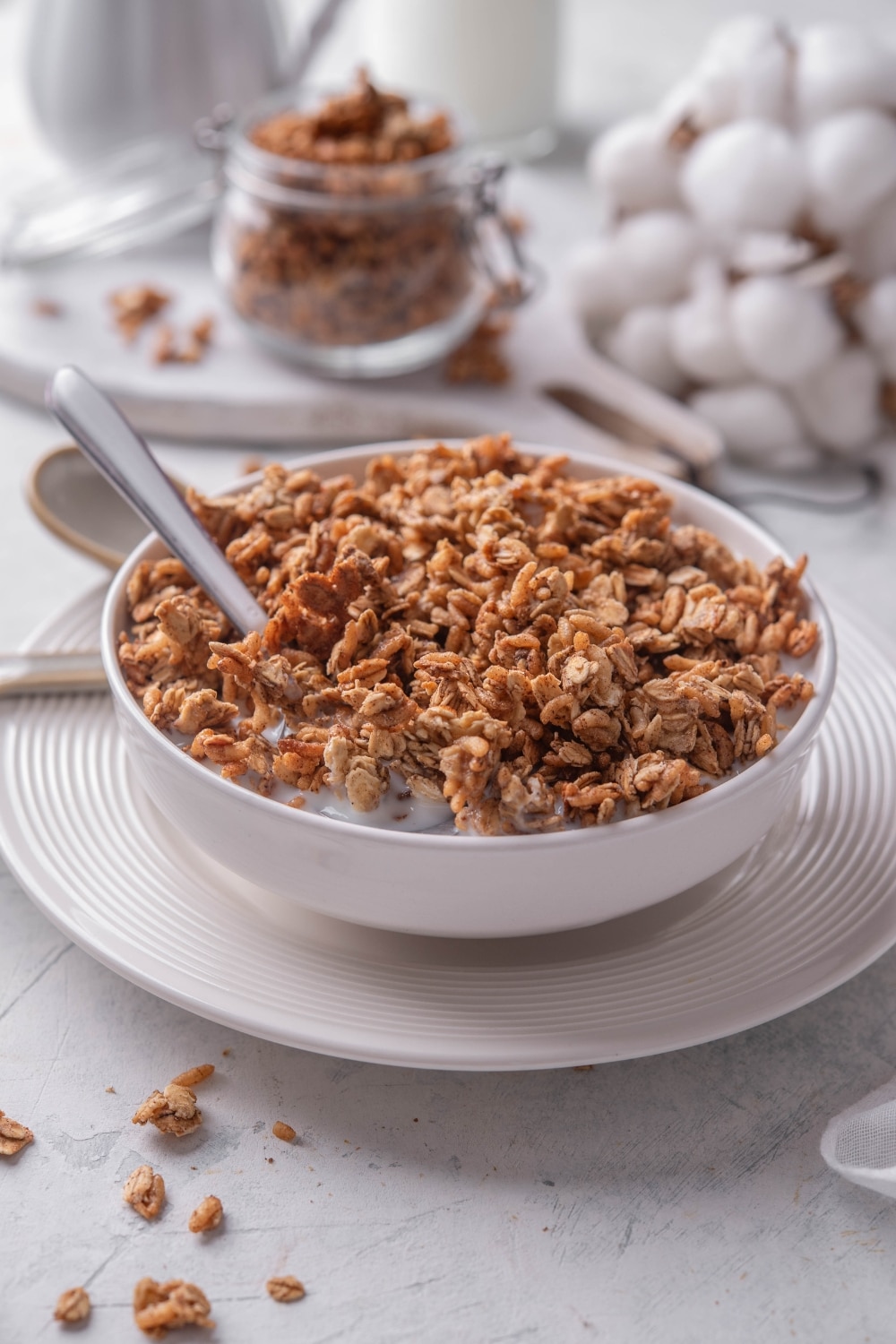 A white shallow bowl filled with granola and milk. There is a spoon in the bowl and the bowl is on a white plate.