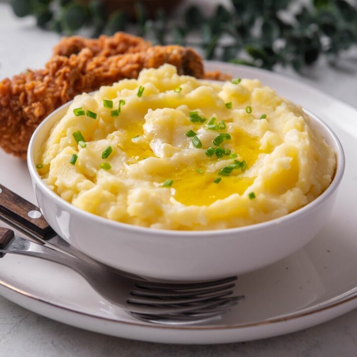 A white ceramic bowl filled with gold mashed potatoes with chives and melted butter on top. The bowl is on a ceramic plate with a piece of friend chicken next to the bowl. There are two forks tucked next to the bowl.
