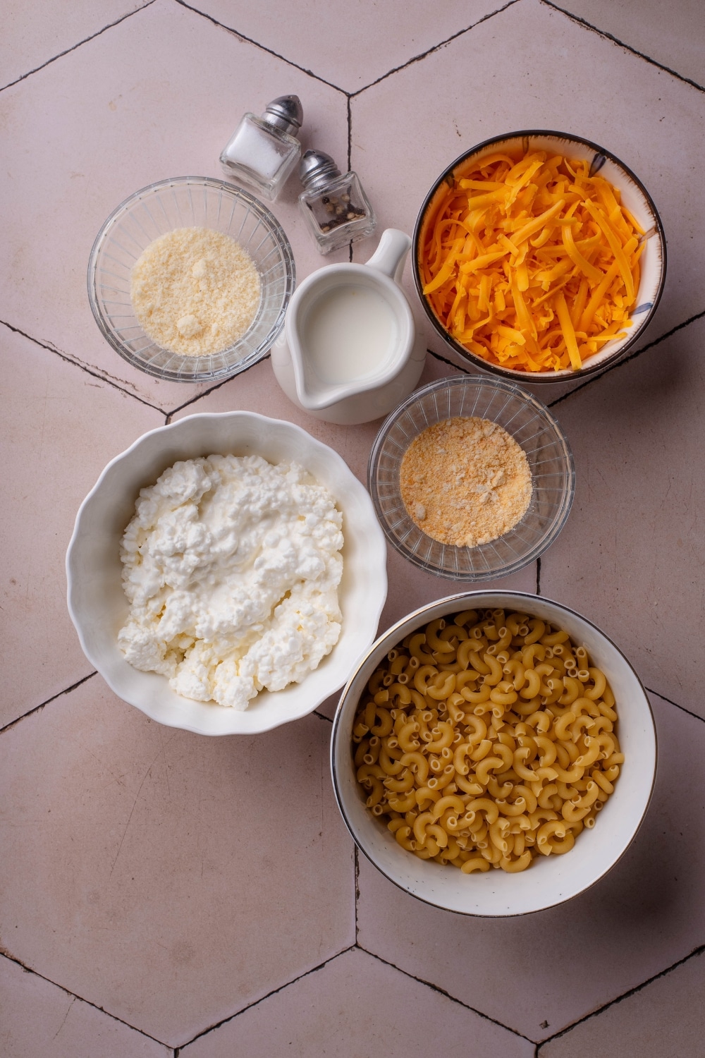 An assortment of ingredients including a pitcher of milk, mini salt and pepper shakers, and bowls of shredded cheese, dried pasta, cottage cheese, and seasonings.