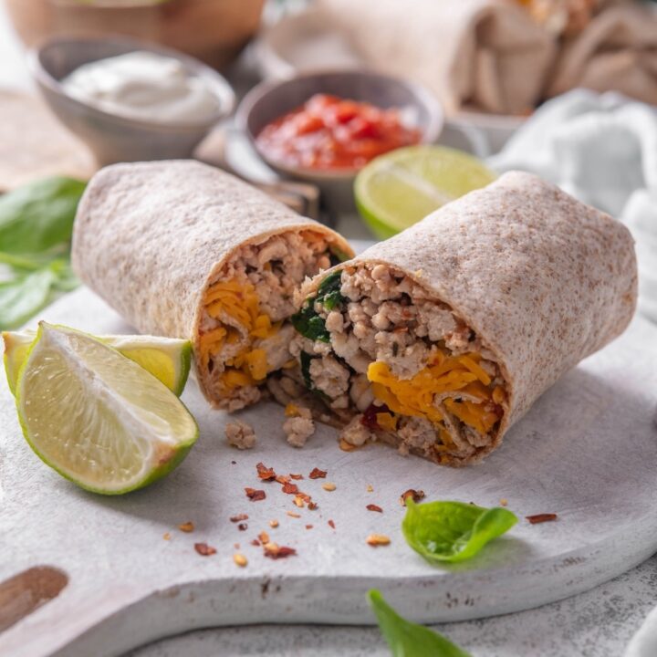 A high protein burrito sliced in half on a wooden server. The burrito has a whole wheat tortilla stuffed with ground turkey, cheese, egg whites, and spinach. There are spinach leaves and red pepper flakes sprinkled on the server.