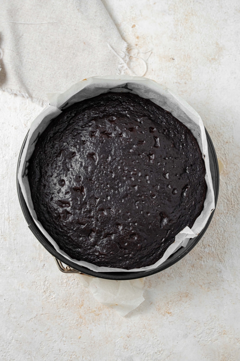 A circular cake pan lined with parchment paper filled with a freshly baked chocolate cake.