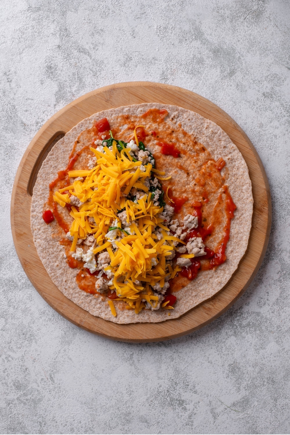 An overhead shot of a whole wheat tortilla with salsa, ground turkey, spinach, and shredded cheese layered on top. The tortilla is on a wooden tray.