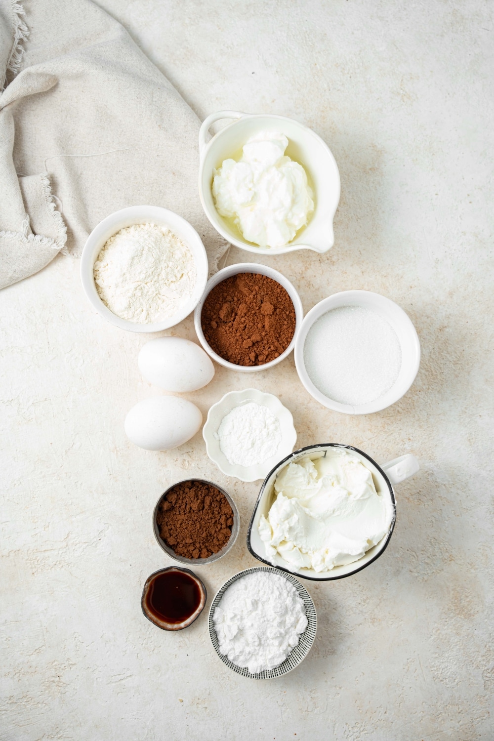 Overhead view of an assortment of ingredients including two eggs and bowls of flour, cocoa powder, vanilla extract, powdered sugar, and yogurt.
