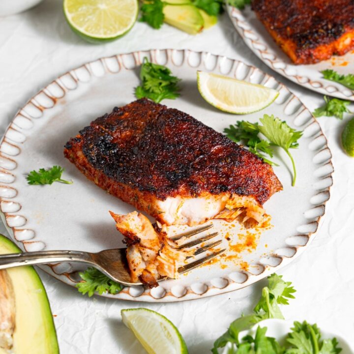 A ceramic plate with blackened mahi mahi with a fork taking a bite of fish. The plate is garnished with parsley and lime. There is a second plate of blackened mahi mahi and additional garnishes next to the plate.