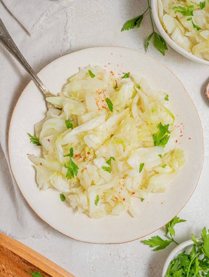 An overhead shot of a white plate with steamed cabbage on a marble counter. There is a fork digging into the cabbage. There is a cloth napkin under the plate with an additional bowl of steamed cabbage and another bowl of fresh parsley off to the side.