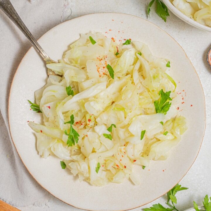 An overhead shot of a white plate with steamed cabbage on a marble counter. There is a fork digging into the cabbage. There is a cloth napkin under the plate with an additional bowl of steamed cabbage and another bowl of fresh parsley off to the side.
