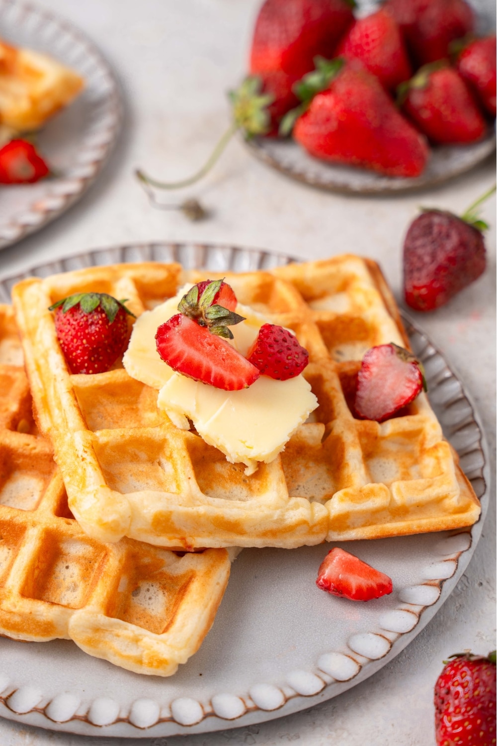 Two cottage cheese waffles on a ceramic plate topped with butter and sliced strawberries. There is a plate of strawberries in the background.