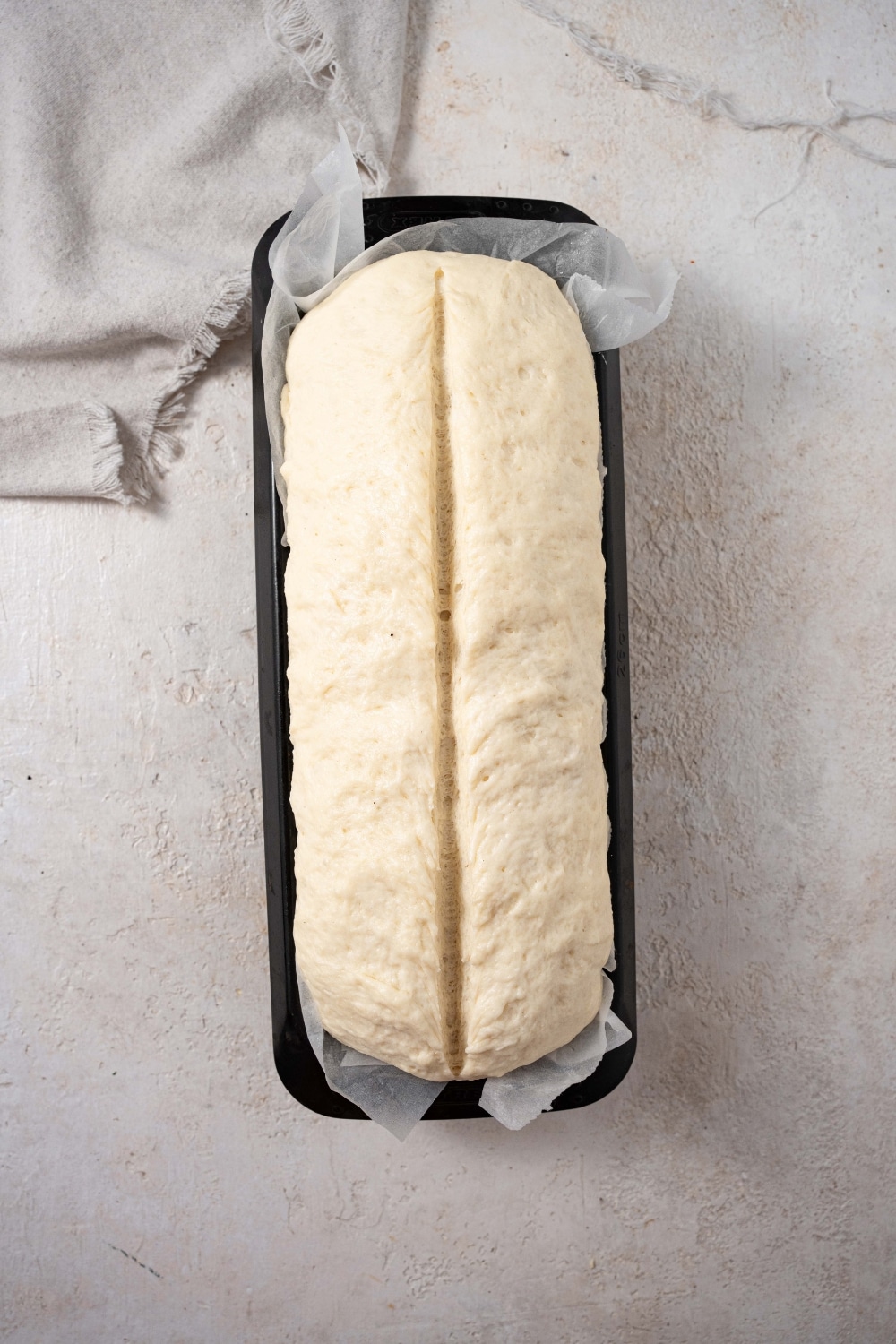 Bread dough scored down the middle in a loaf pan lined with parchment paper.