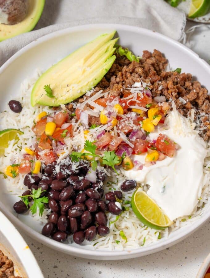 A taco bowl with taco meat, sliced avocado, rice, veggies, beans, and cottage cheese sauce.