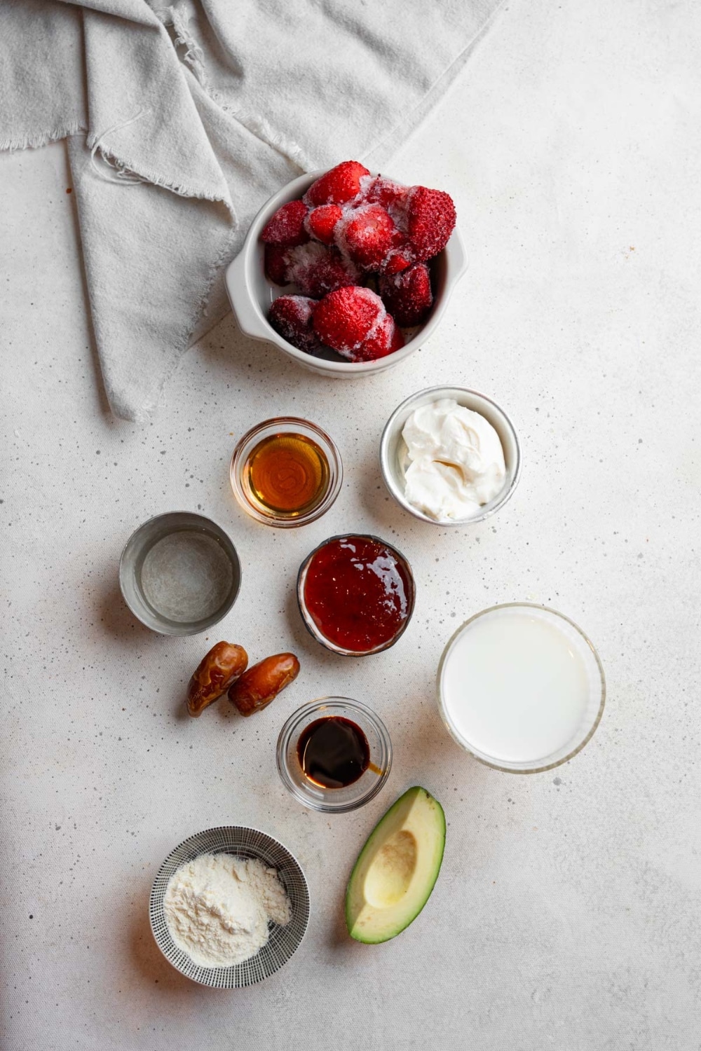 An overhead shot of several bowls in various sizes containing ingredients to make Hailey Bieber smoothie recipe including frozen strawberries, avocado, dates, collagen powder, coconut cream, hyaluronic acid liquid, maple syrup, and vanilla.