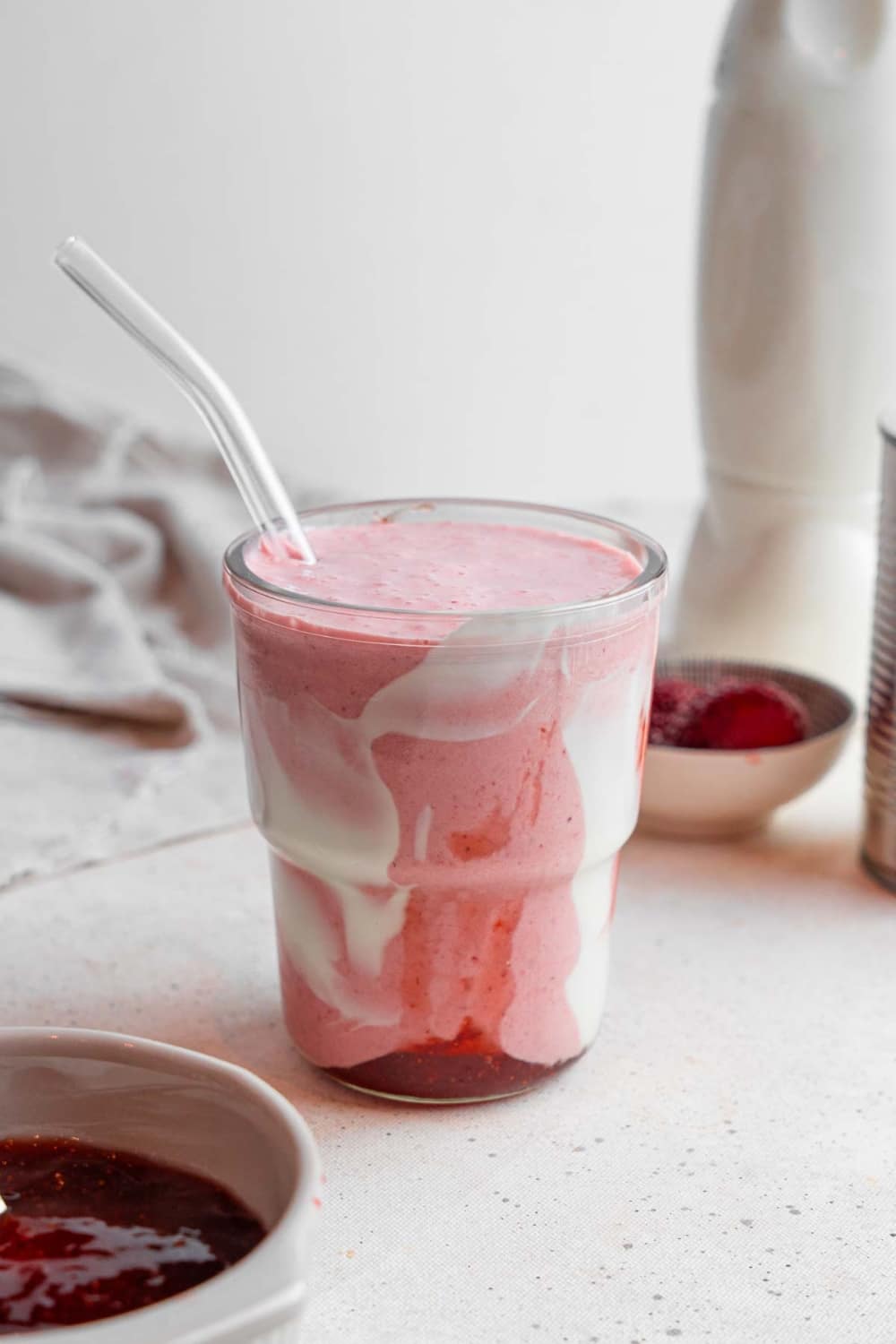 A Hailey Bieber smoothie with a glass straw on a marble counter. The glass is decorated with coconut cream and strawberry jam and the counter has a bowl of jam and bowl of frozen strawberries in the background.