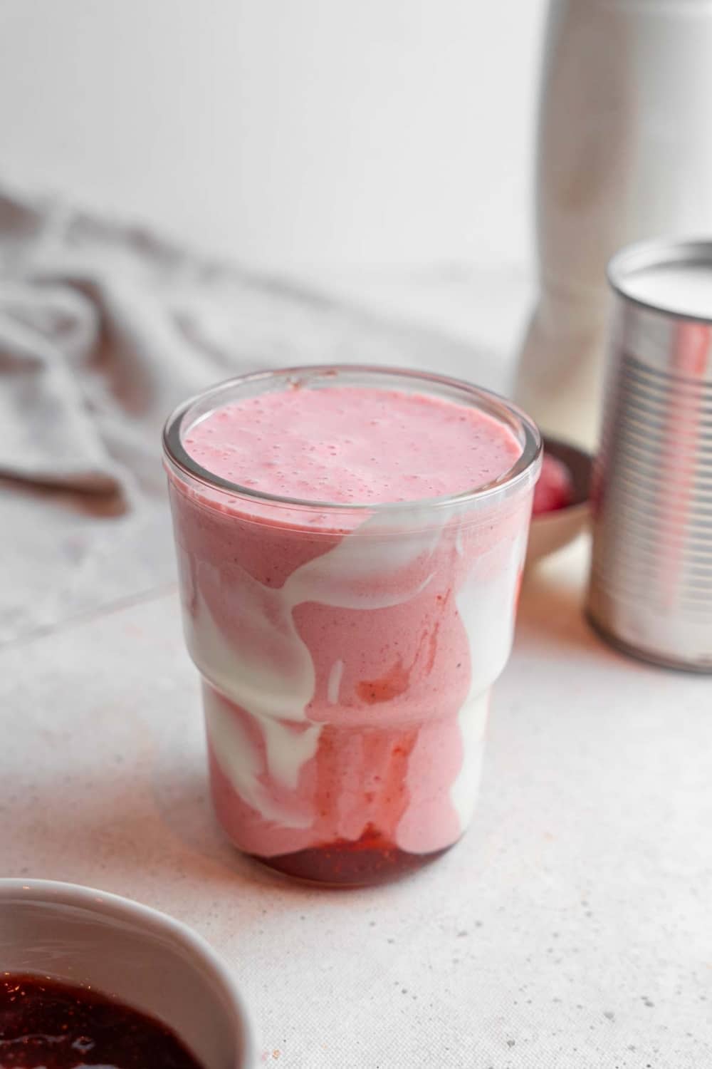 A Hailey Bieber smoothie on a marble counter. The glass is decorated with coconut cream and strawberry jam and the counter has a bowl of jam and frozen strawberries in the background.